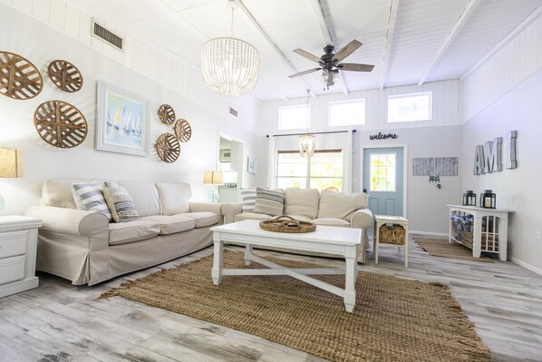 A spacious living room with a beige sofa, a white coffee table, and a chandelier. There are decorative wall art pieces and a 'welcome' sign on the wall.