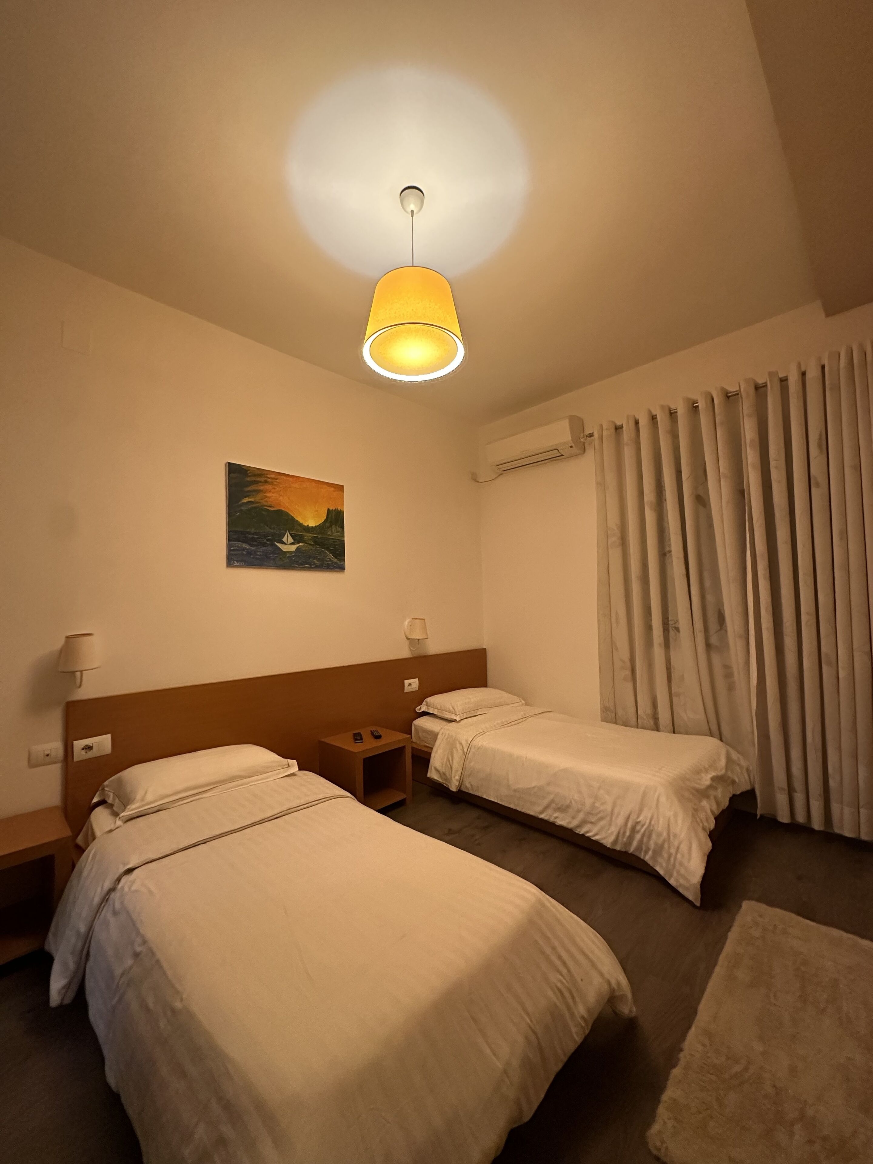 Ein Hotelzimmer mit zwei Einzelbetten, einem Nachttisch, einer Lampe, einer Klimaanlage, einem Bild an der Wand und einem Vorhang.