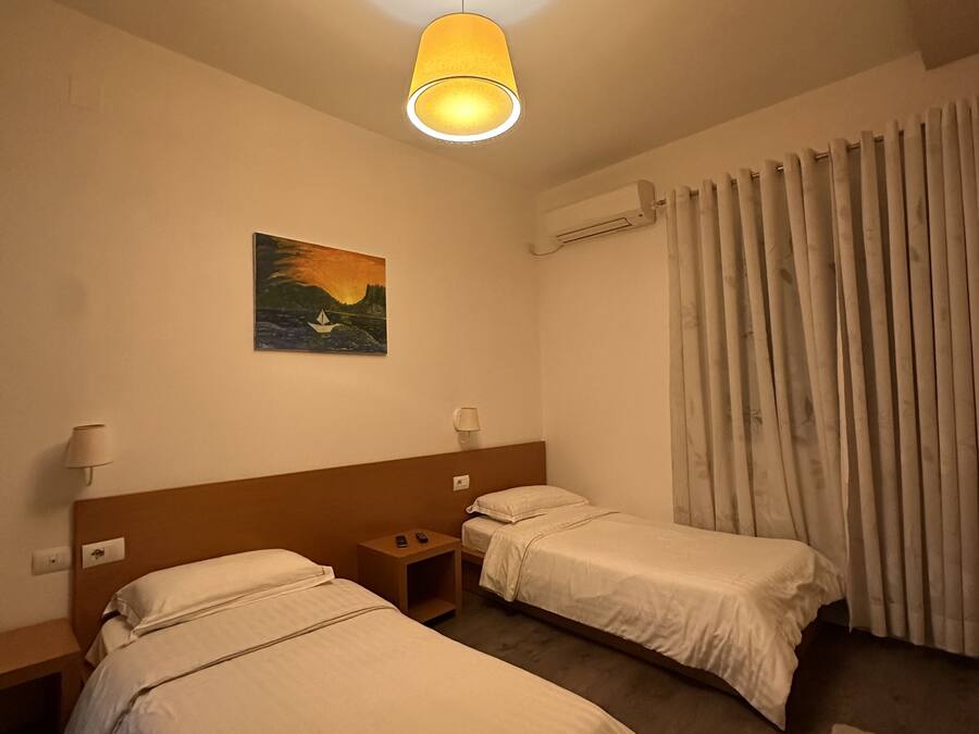 Ein Hotelzimmer mit zwei Einzelbetten, einem Nachttisch, einer Lampe, einer Klimaanlage, einem Bild an der Wand und einem Vorhang.