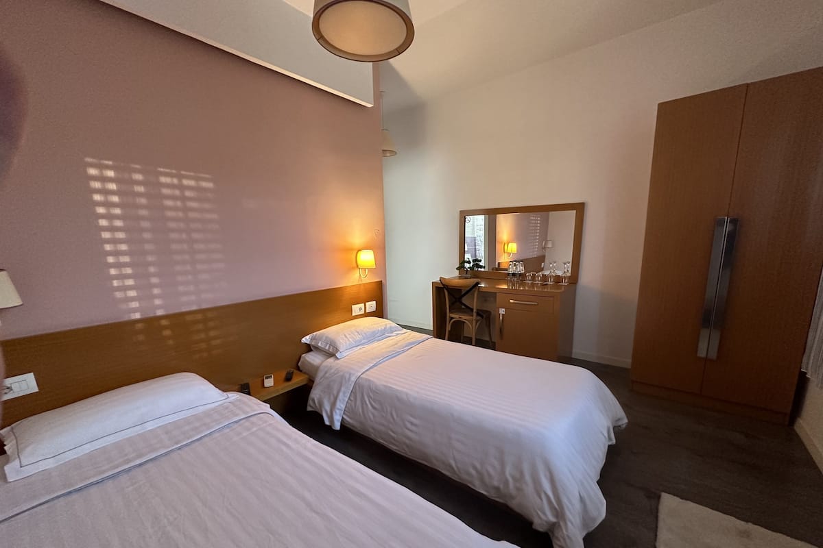 Quarto de hotel com duas camas, uma escrivaninha, um espelho e um guarda-roupa.