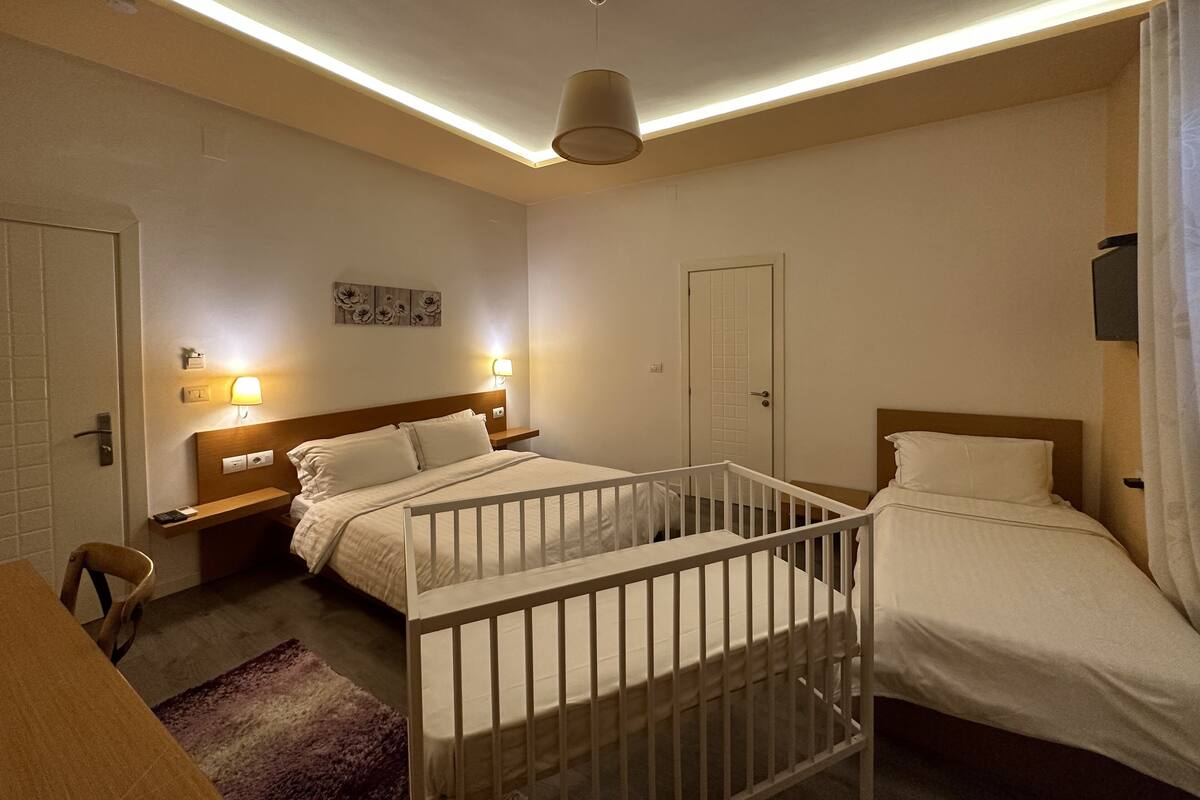 Quarto de hotel com duas camas, um berço, uma mesa de cabeceira, uma luminária fixada na parede e uma porta.