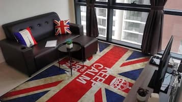 A modern living room with a sofa, a small round table, and a television. A Union Jack-themed rug is on the floor, and the room has large windows with curtains.