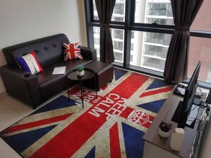 A modern living room with a sofa, a small round table, and a television. A Union Jack-themed rug is on the floor, and the room has large windows with curtains.