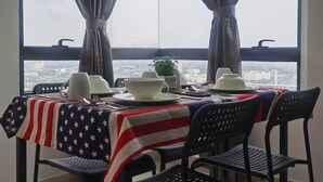 Une table à manger, dressée avec des assiettes, des bols et des couverts, recouverte d’une nappe aux couleurs du drapeau américain, placée devant une fenêtre ornée de rideaux et donnant sur un paysage urbain.