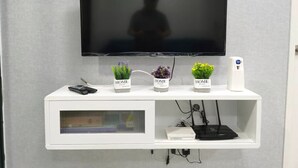 Un téléviseur à écran plat fixé à un mur blanc, avec trois petites plantes en pot étiquetées « MAISON » sur une étagère blanche située en dessous.