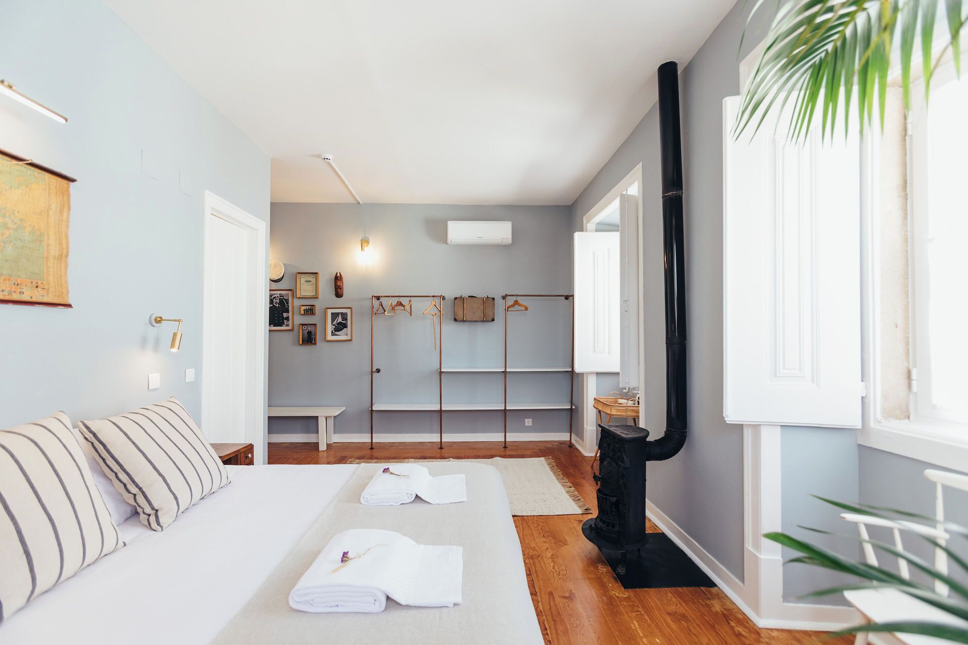 A room with a bed, towels, a coat rack, a black heater, and a window with a plant.