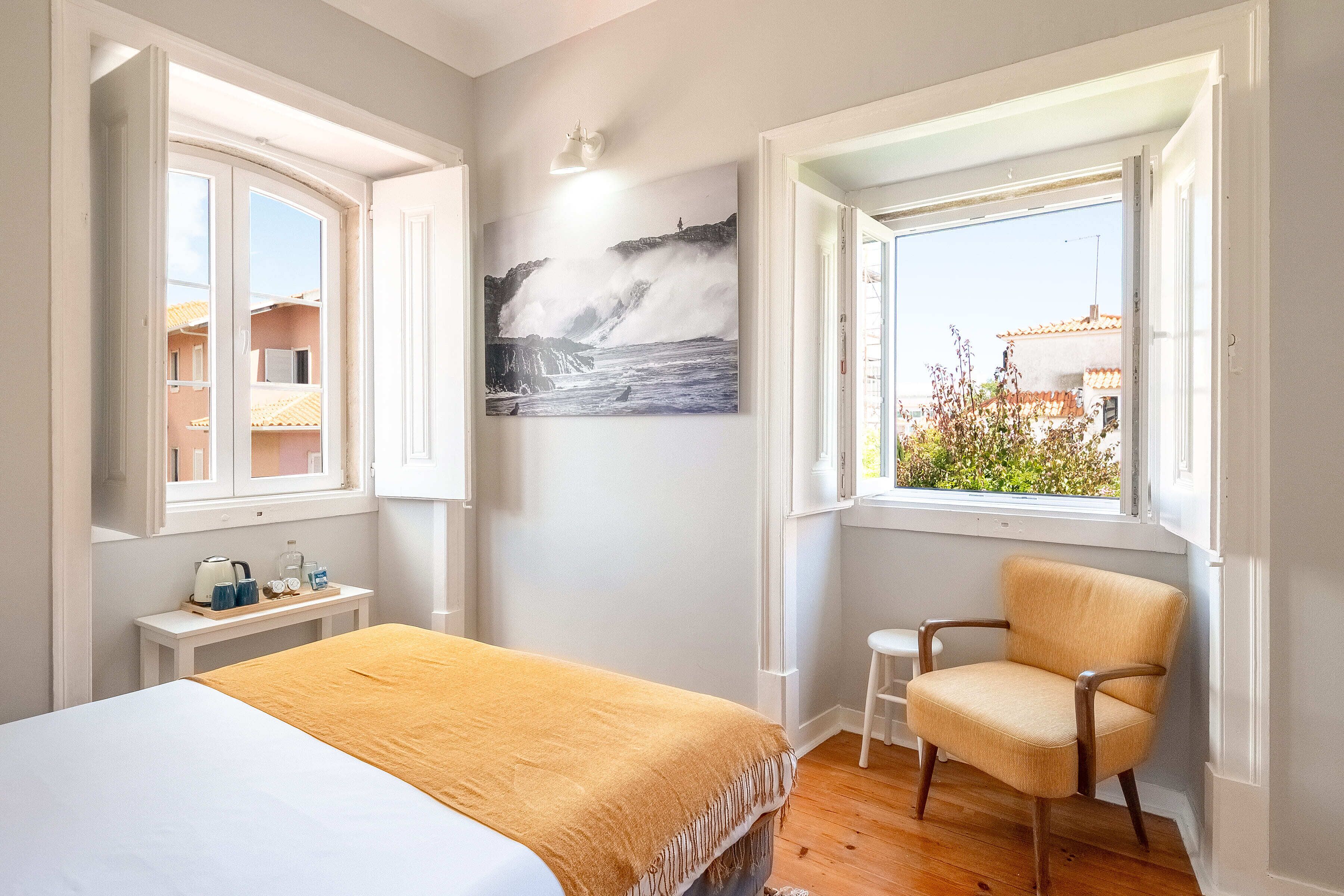 A bedroom with a bed, a chair, a small table, a painting, and a window with a view.