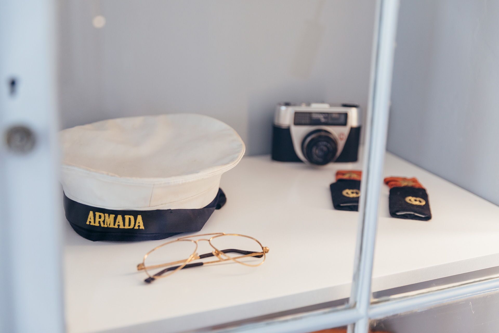 A display case containing a navy blue and white cap labeled "ARMADA," a vintage camera, a pair of eyeglasses, and two black patches with yellow emblems.