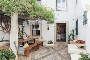 A courtyard with a wooden table, benches, a pergola with vines, a tiled floor, and a doorway leading to a kitchen. - Legasea Cascais (Cascais)
