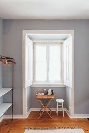 A corner room with a window, a wicker table with kitchen items, a white chair, and a wooden floor. - Legasea Cascais (Cascais)