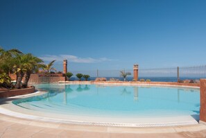 Piscina azul cristalina con plataforma de azulejos, rodeada de palmeras y una valla, con vista a una extensión de agua.