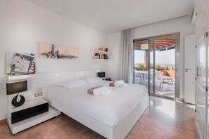Una habitación de hotel moderna con una cama grande, un mueble para televisión, un cuadro de un paisaje urbano y un balcón con mobiliario exterior.