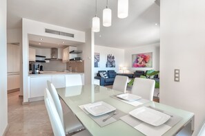 A modern kitchen with white cabinets, a central island, and a dining area with a glass table set for two. - Bahia - Alzaro 1.3 DELUXE SEA VIEW & JACUZZI 3B (Arona)