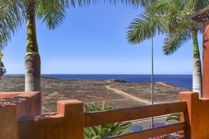 A view from a wooden balcony overlooking a coastal landscape with palm trees and a clear blue sky.