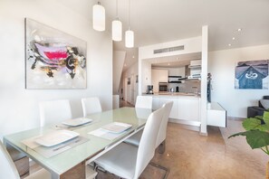 A modern dining area with a glass table, white chairs, and a kitchen in the background. - Bahia - Alzaro 1.3 DELUXE SEA VIEW & JACUZZI 3B (Arona)
