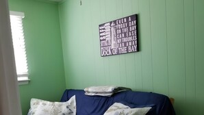 Une pièce aux murs verts, avec un lit en bois recouvert d’une literie bleue et agrémenté d’oreillers à motifs floraux, un ventilateur de plafond et une citation inspirante sur le mur.