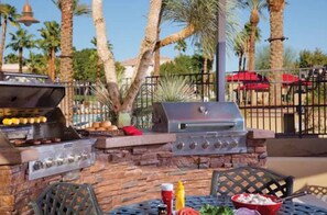 Area barbecue esterna con muro in pietra, tavolo e sedie, e vista su palme e un edificio sullo sfondo.