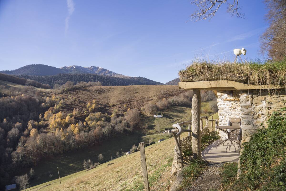 A stone bridge with a table and chairs, overlooking a valley with trees and a mountain in the background.