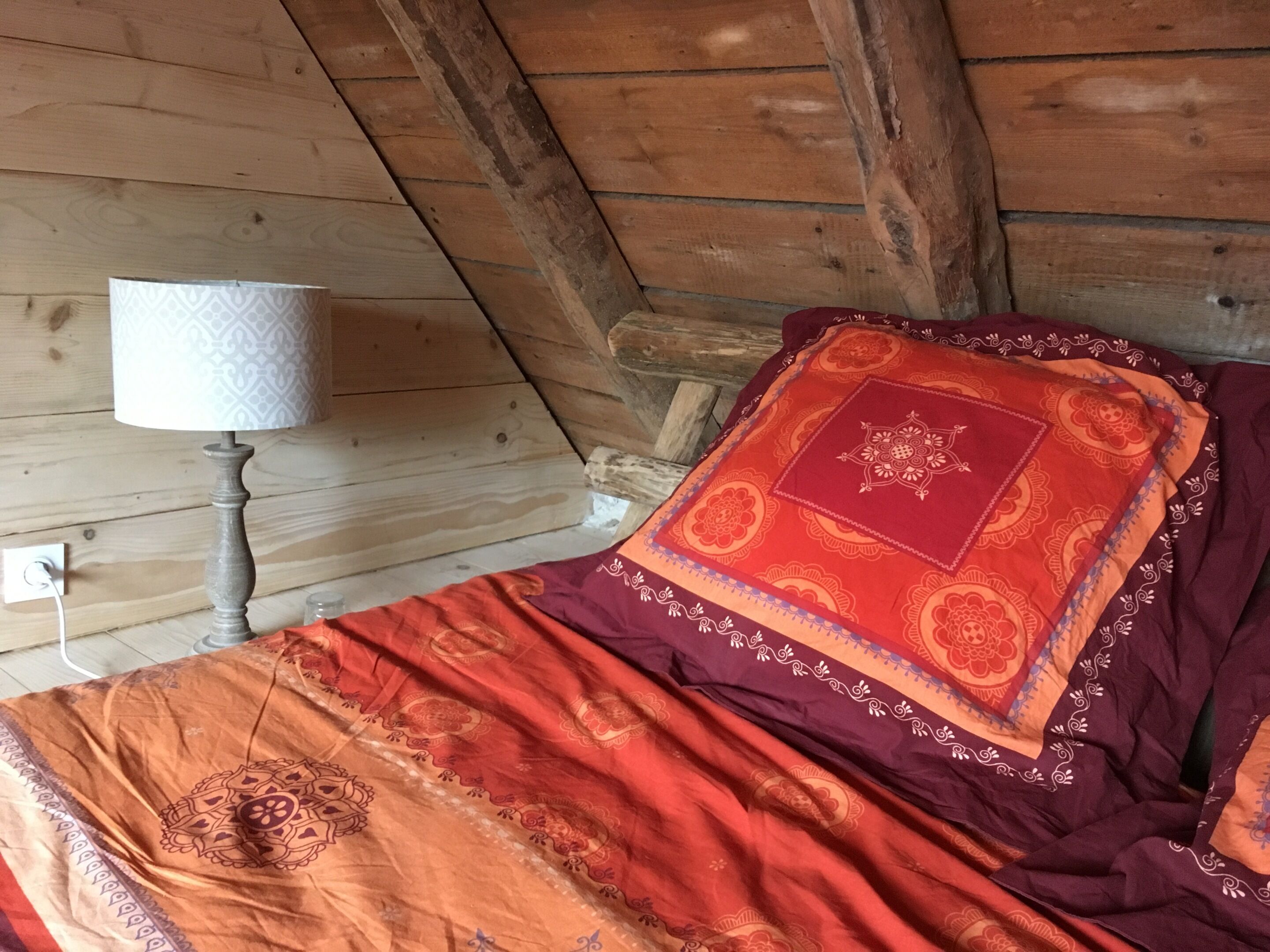 Techo de madera con diseño inclinado, una cama con manta estampada y almohada decorativa, y una lámpara de noche.