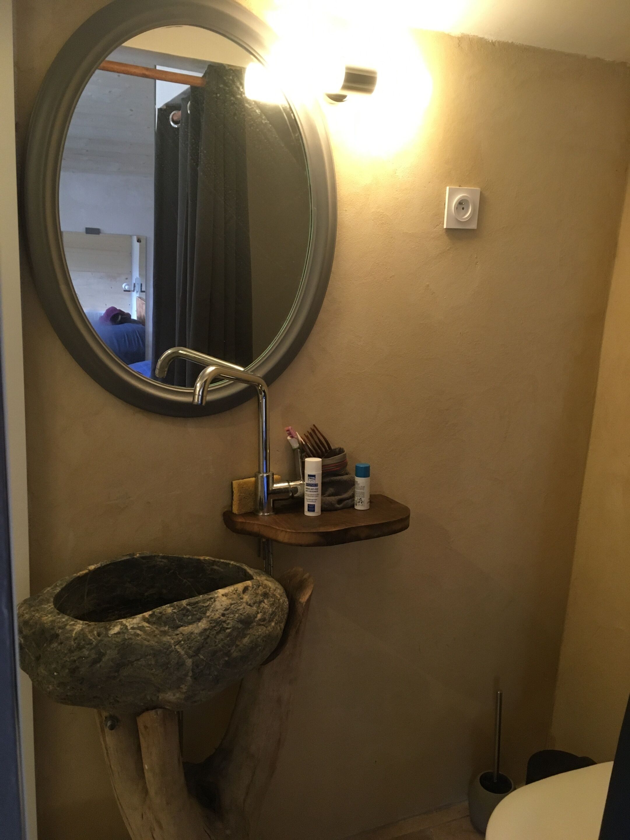 A bathroom with a round mirror, a wooden shelf with toiletries, a stone sink, and a toilet.