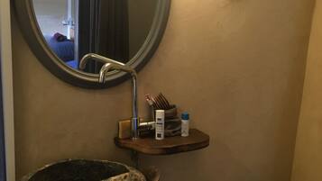 A bathroom with a round mirror, a wooden shelf with toiletries, a stone sink, and a toilet.