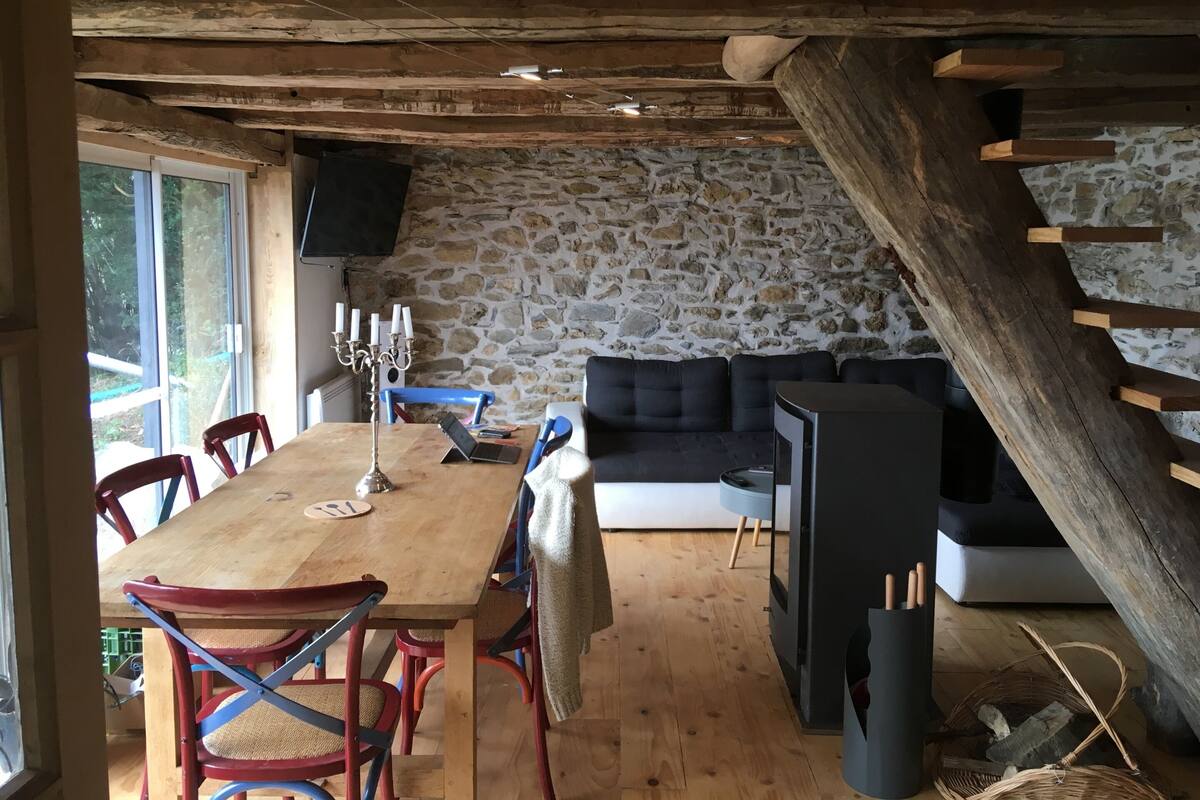 A rustic cabin interior with a stone wall, wooden ceiling beams, a dining table with chairs, a sofa, and a fireplace.
