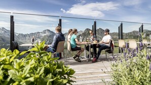 Un groupe de personnes est assis à une table en plein air et déjeune, avec une vue sur les montagnes.