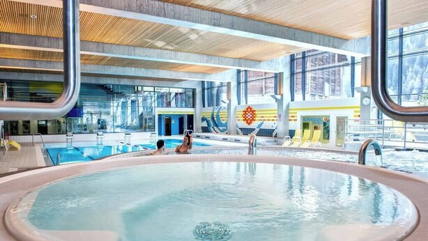 Une piscine intérieure avec un jacuzzi, entourée de chaises longues et offrant une vue sur l’extérieur.