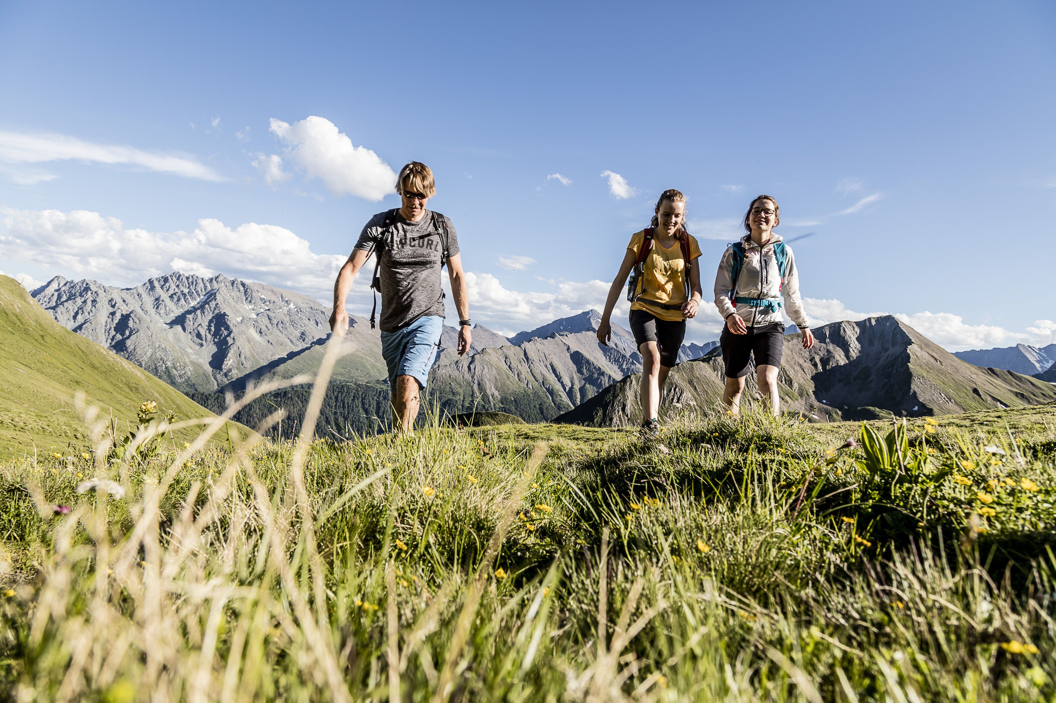 Tre persone che fanno un'escursione su un sentiero montano erboso, con montagne sullo sfondo.