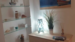 A room with a wooden dresser, glass shelves with decorative items, a potted plant, and a painting of a boat on water. - Stella Marina - Stella Studio (Polignano a Mare)