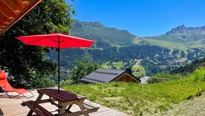 A wooden deck with a picnic table and a red umbrella, overlooking a mountainous landscape with a small building in the distance. - Homerez - Chalet in Les Avanchers-Valmorel (Les Avanchers-Valmorel)