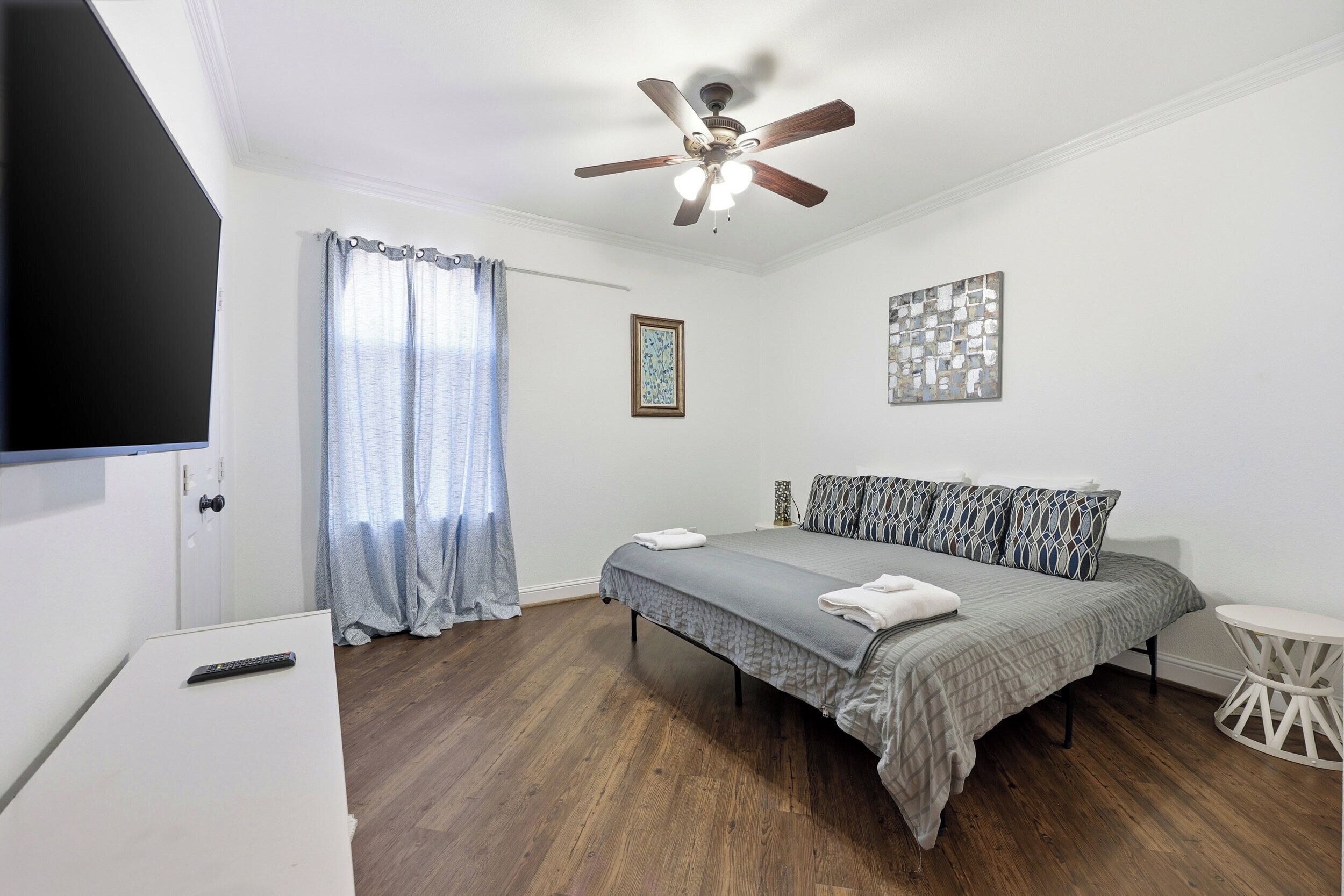A bedroom with a bed, a ceiling fan, a TV, a window with curtains, and a small table.
