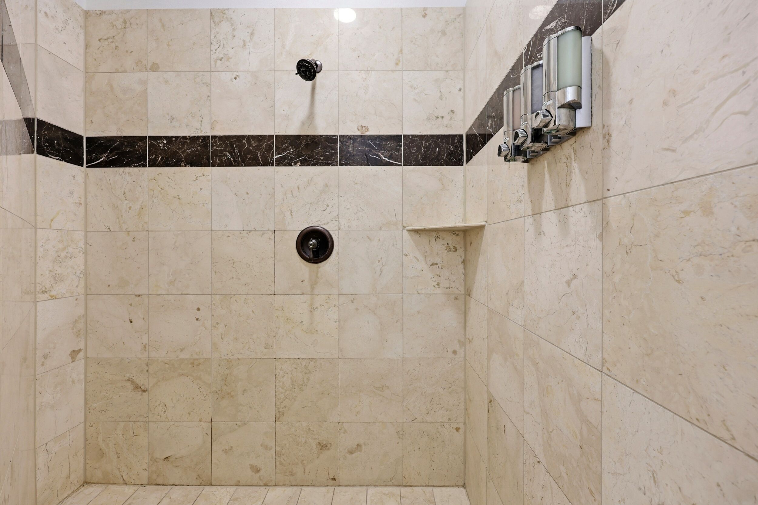 Ein gefliester Duschbereich mit einem schwarzen und beigen Fliesendesign, einer Duschbrause und einem an der Wand montierten Warmwasserbereiter.