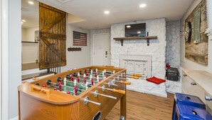 A room with a foosball table, a stone fireplace, a mounted television, and a dartboard.