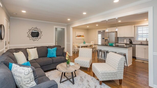 A living room with a dark grey sofa, patterned armchairs, and a round coffee table. A kitchen is visible in the background with white cabinets and stainless steel appliances.