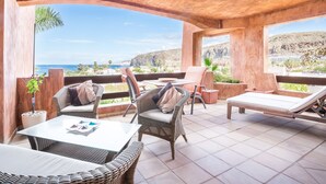 Un espacio de estar al aire libre cubierto con mobiliario de mimbre, una mesa de vidrio y vistas al mar y las colinas.