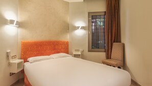 Une chambre d’hôtel avec un grand lit, deux tables de chevet, une chaise, une fenêtre avec des rideaux et des lampes fixées au mur.