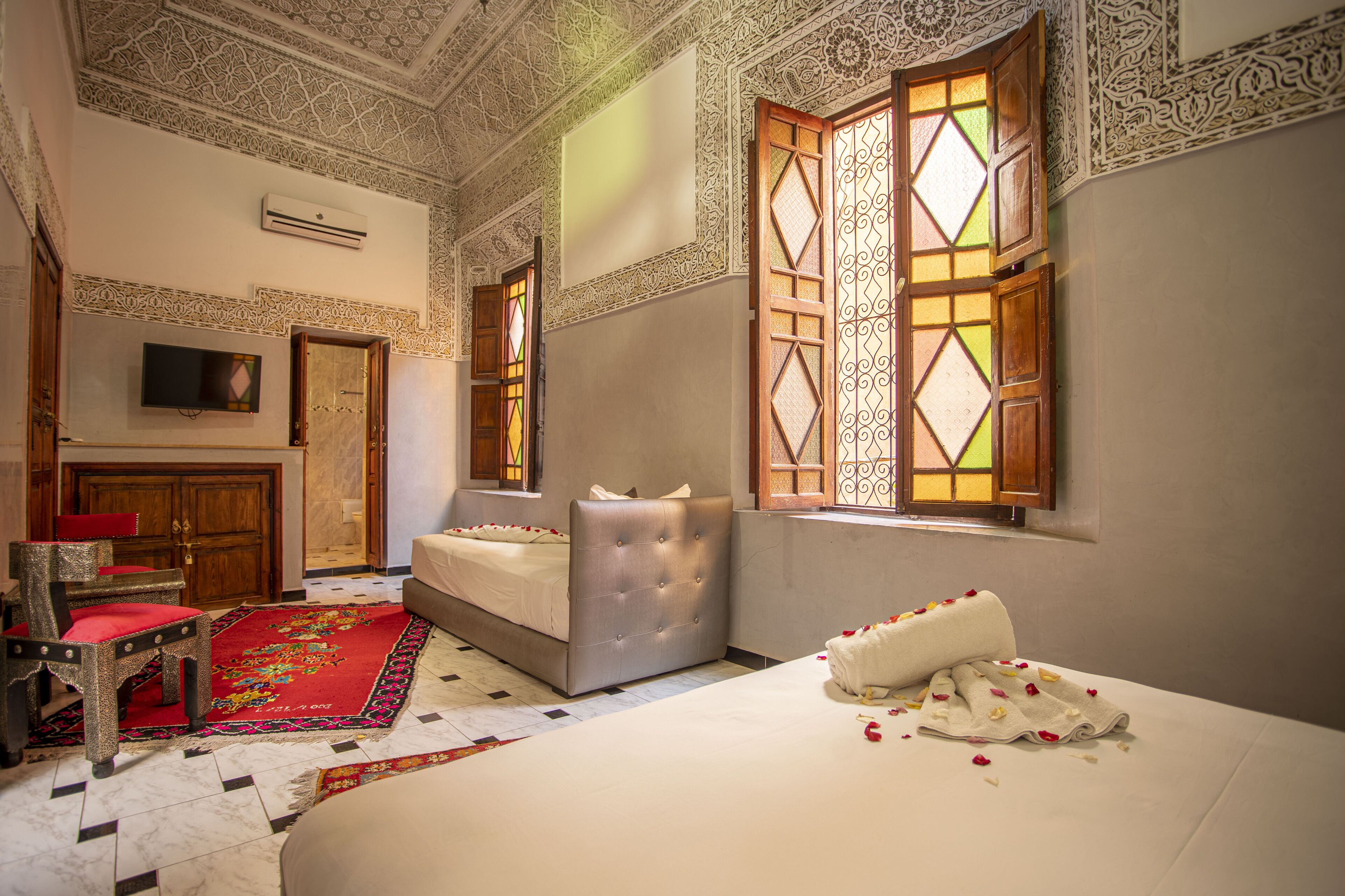A hotel room with a bed, a chair, a TV, and a window with intricate patterns.