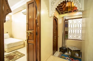 Una camera d'albergo con un letto, una porta in legno, un tavolino e una finestra con inferriate decorative.