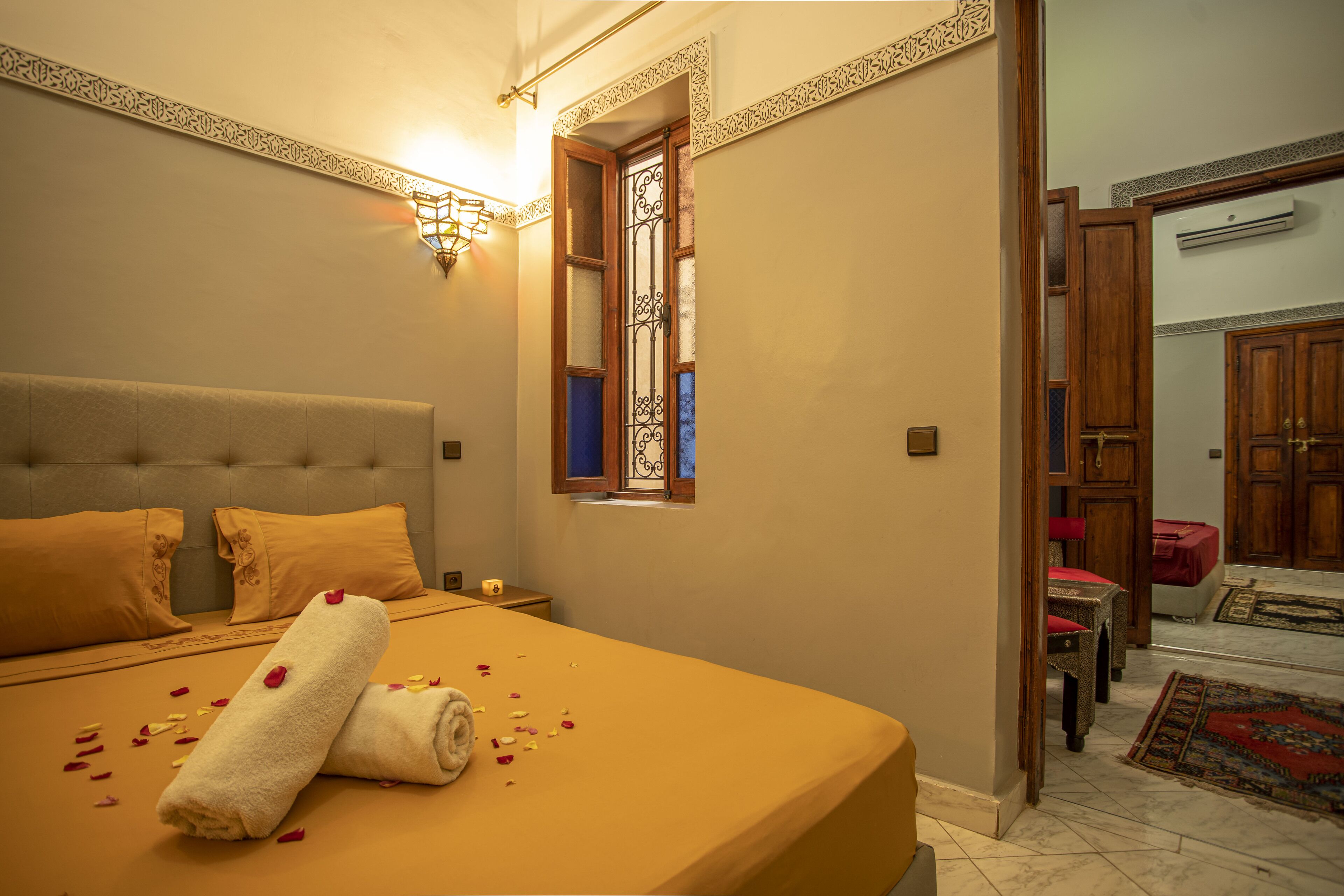 Habitación de hotel con una cama grande, una mesita pequeña, un ventanal con rejas decorativas y vistas al pasillo.