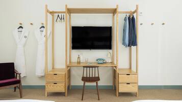 A room with a wooden desk, a chair, a TV, and a bed. There are towels and robes hanging on the wall.