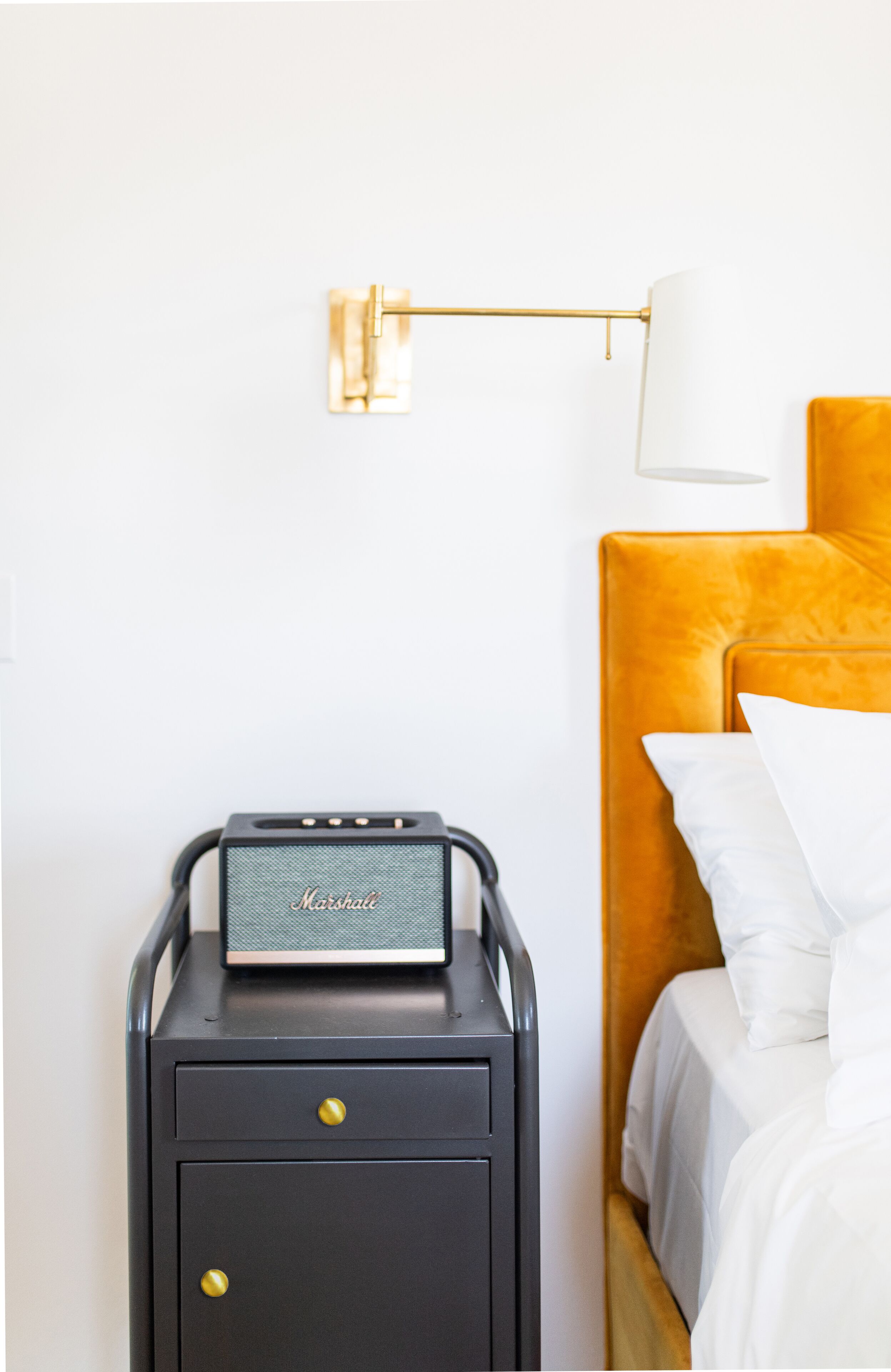 Une chambre compacte et moderne, avec un lit, une petite table de chevet noire sur laquelle se trouve un haut-parleur Marshall, et une lampe dorée fixée au mur.