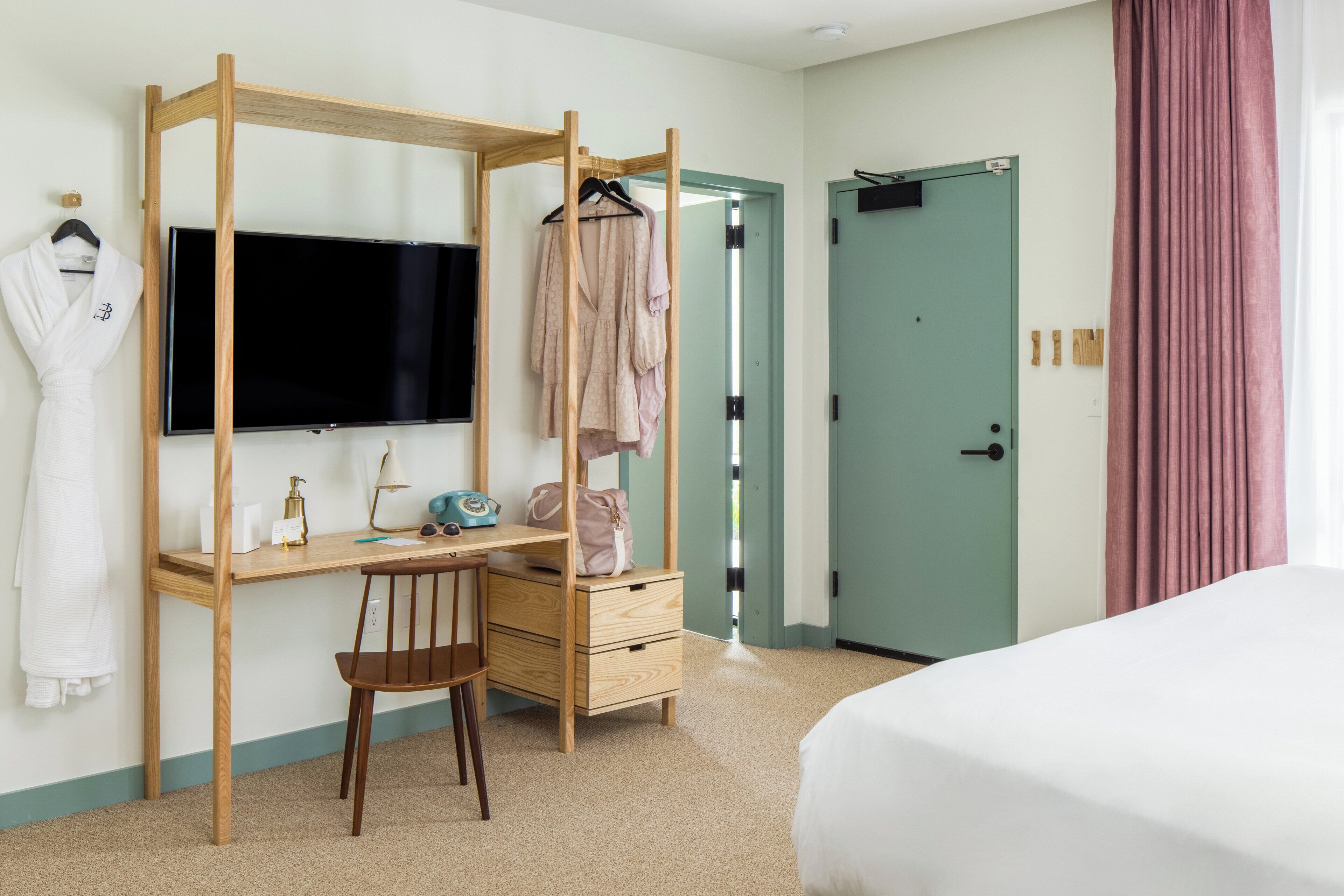 A hotel room with a wooden desk, a chair, a bed, a TV, and a door.