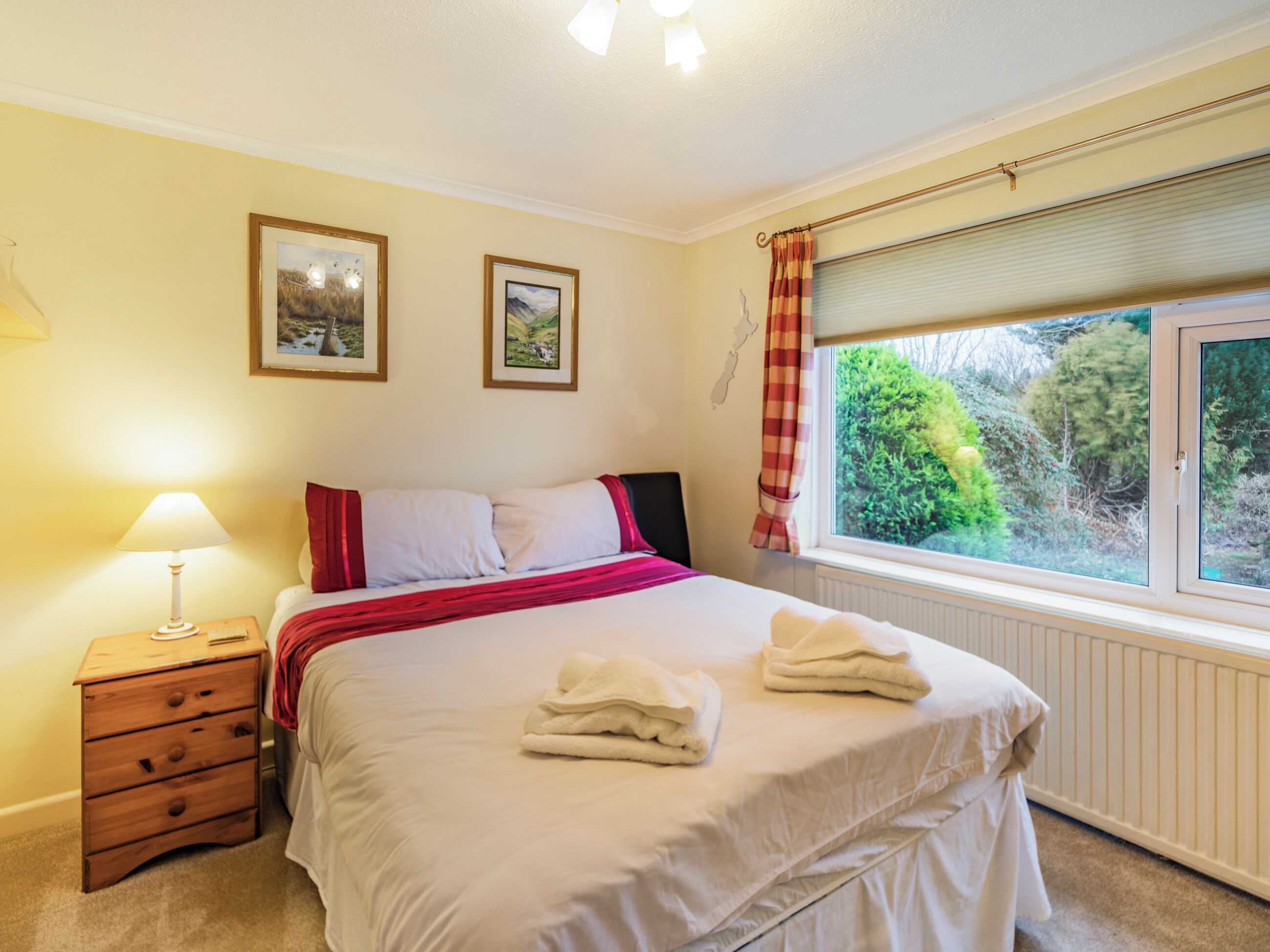 A bedroom with a bed, a nightstand, a lamp, two framed pictures on the wall, and a window with curtains.
