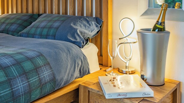 Een netjes opgemaakt bed met ruitbeddengoed, een houten hoofdbord, een nachtkastje met een champagnefles, twee wijnglazen en een menukaart.