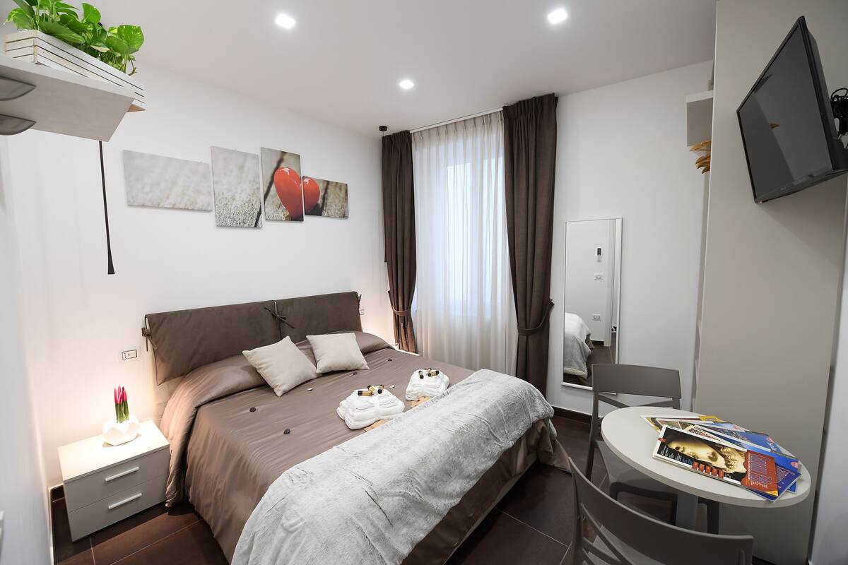 A modern hotel room with a large bed, a bedside table with a lamp, a small table with magazines, and a TV mounted on the wall.