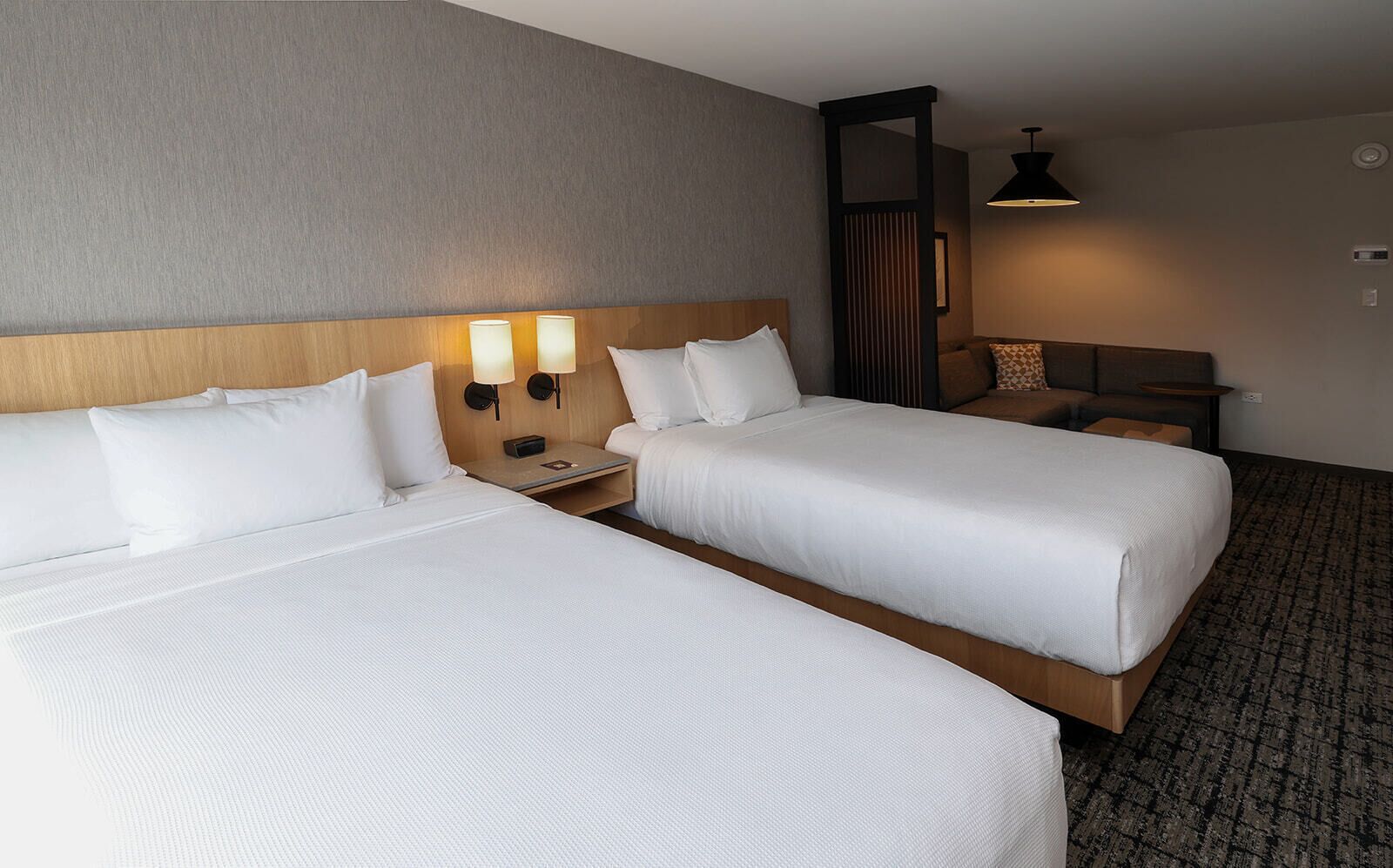 A hotel room with two single beds, a nightstand with a lamp, a wooden headboard, and a small seating area with a table and chair.