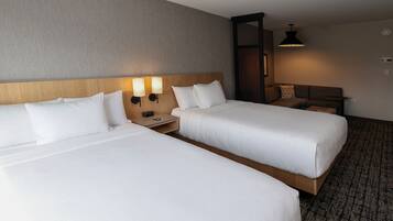 A hotel room with two single beds, a nightstand with a lamp, a wooden headboard, and a small seating area with a table and chair.