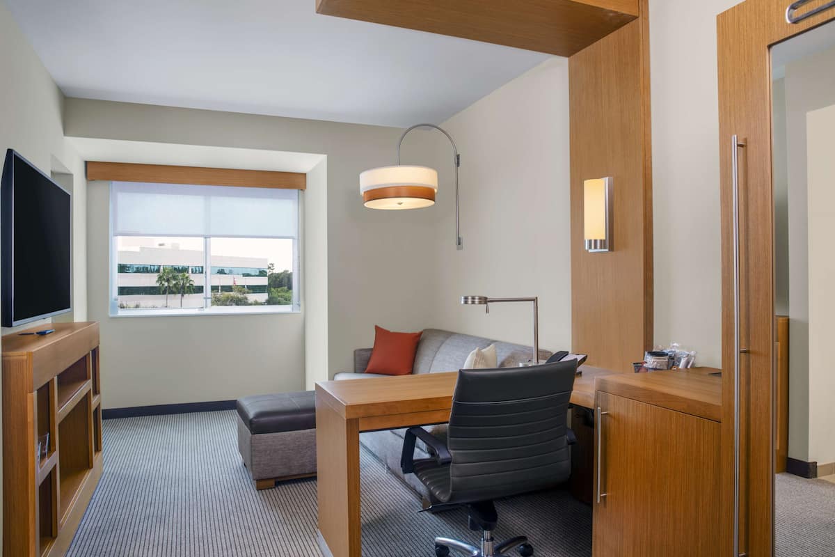 A modern hotel room with a wooden desk, a comfortable sofa, a flat-screen TV, and a large window offering a view of the outside.