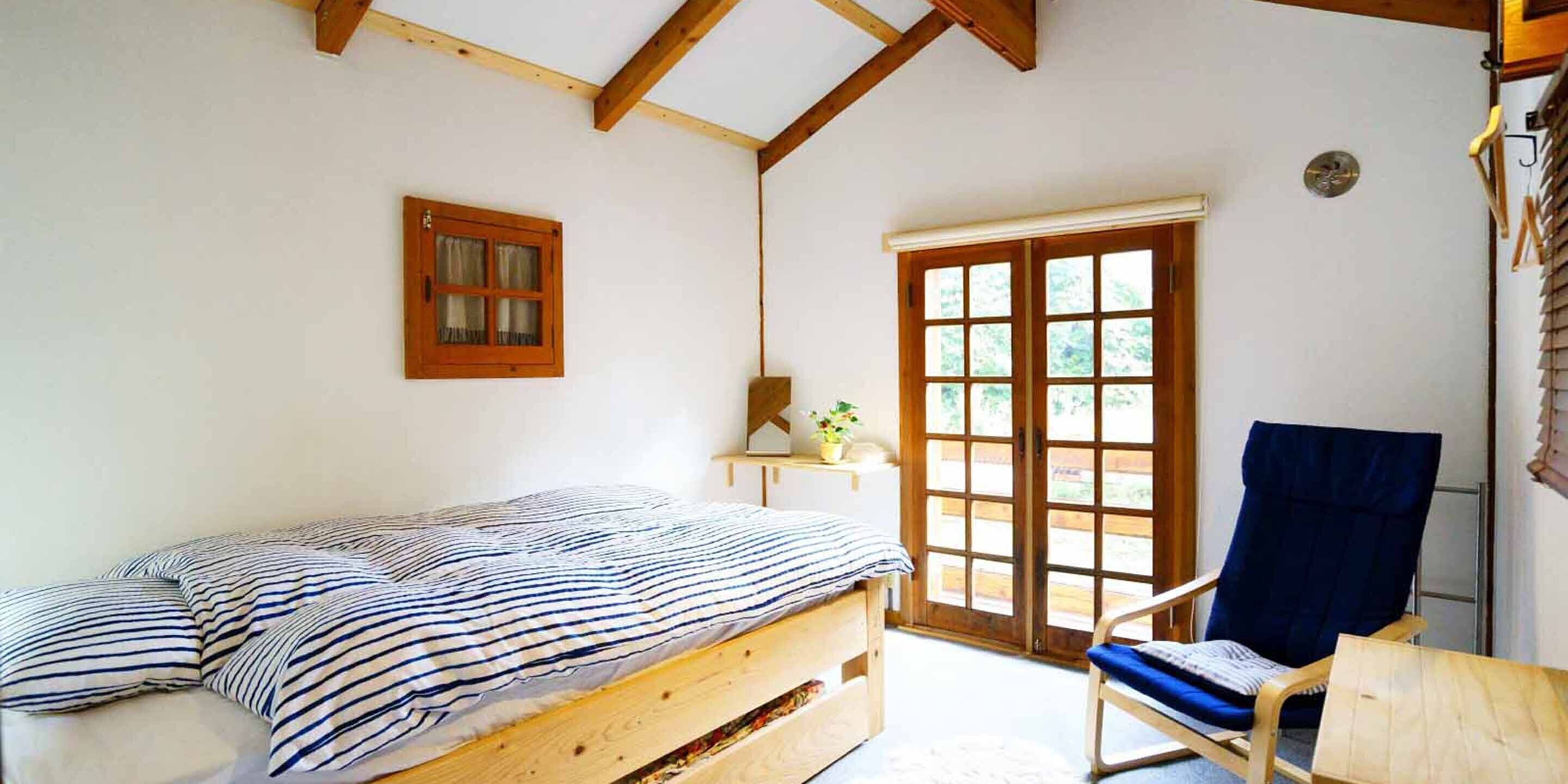A room with a wooden bed, a chair, a window with a view, and a door.